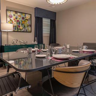 A modern dining room with a large table set for a meal, featuring elegant chairs, artwork on the wall, and warm lighting.