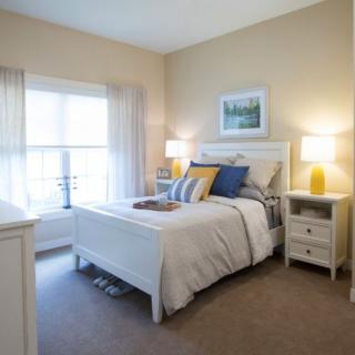 A cozy bedroom with light-colored walls, featuring a bed with decorative pillows, nightstands with lamps, and natural light streaming through the window.