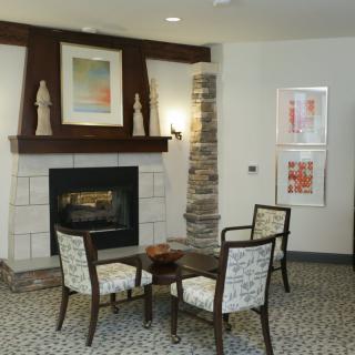 A cozy living room setting with a fireplace, chairs, and artwork on the walls.