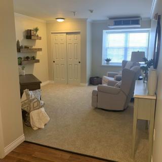 A cozy living room with carpeted flooring, featuring comfortable seating and natural light streaming through a large window.