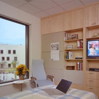 A cozy hospital room with a view, featuring a bed, chair, bookshelf, and television.
