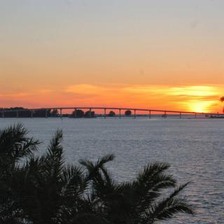 A beautiful sunset over a bridge, with palm trees in the foreground and a calm body of water.