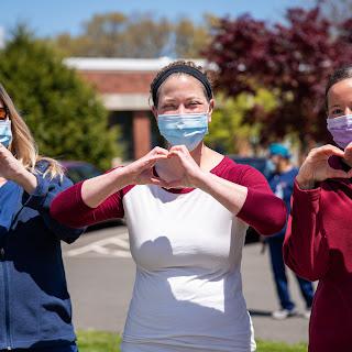 Three individuals show their support and solidarity by forming heart shapes with their hands while wearing masks outdoors.