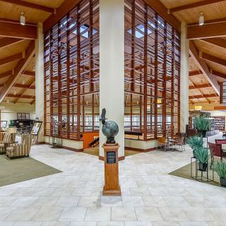 Modern library interior with wooden beams and large glass windows, featuring a central statue.