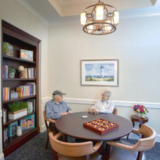 Elderly individuals sitting in a cozy room with a chess board on the table and a bookshelf nearby.