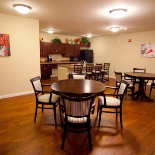 A cozy dining area with wooden flooring, featuring multiple tables and chairs adjacent to a kitchen.