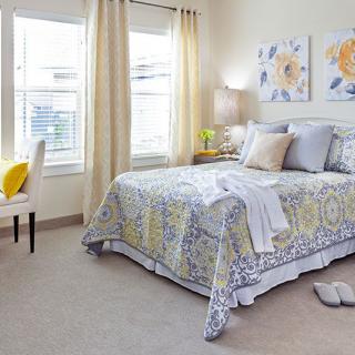 A cozy and well-lit bedroom featuring a patterned bedspread, two sunny windows, and artwork on the walls.