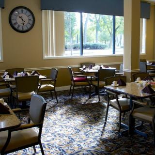 An empty dining room with tables and chairs, featuring a large clock on the wall and natural light from windows.