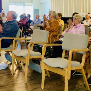 A group of seniors seated indoors, applauding during an event.