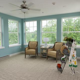 A bright sunroom with large windows, featuring two chairs, a small table, and an easel.