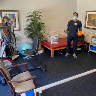 A healthcare worker wearing a mask in a physical therapy room with various exercise equipment.