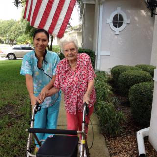 An elderly woman with a walker, accompanied by a caregiver, standing in front of a house with an American flag.