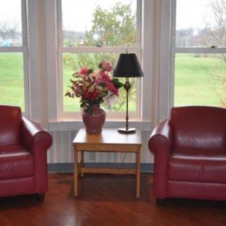 A cozy seating area with two red chairs, a table with flowers, and a lamp in front of large windows overlooking greenery.