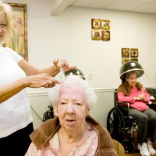 A woman styles the hair of an elderly lady at a salon, with another person seated under a hairdryer in the background.