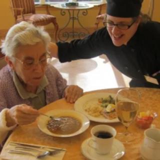 An elderly person enjoys a meal with assistance from a caregiver.