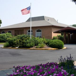 A brick building with an American flag flying in front, surrounded by lush landscaping and colorful flowers.