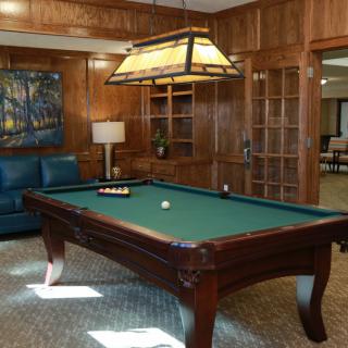 A cozy game room featuring a billiards table with a hanging lamp above it, surrounded by wood-paneled walls and comfortable seating.
