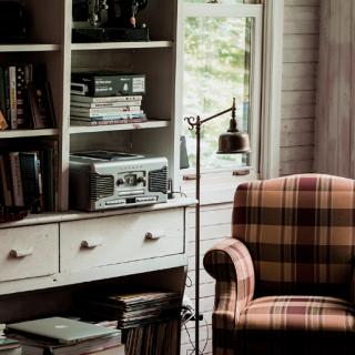 A cozy living room corner with a plaid armchair, a well-stocked bookshelf, and a standing lamp by the window.
