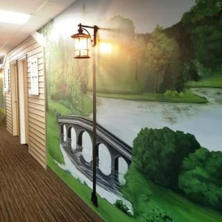 A hallway featuring a scenic mural of a bridge over a river, illuminated by a wall lamp.
