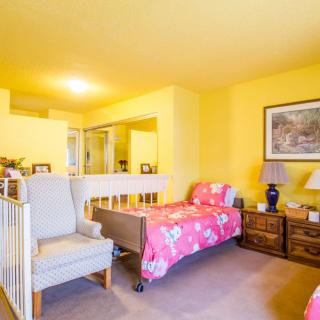 A cozy bedroom with yellow walls, featuring a single bed covered in a pink blanket, an armchair, and a nightstand with a lamp.