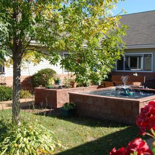 A sunny backyard garden with a central fountain surrounded by brick walls and lush greenery.