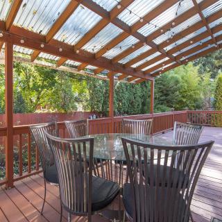 A cozy outdoor patio with a wooden pergola and glass table set amidst lush greenery.