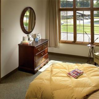 A cozy bedroom featuring a wooden dresser, round mirror, and large window with a view.