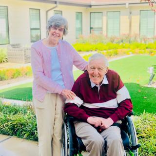 An elderly couple enjoying time together in a garden, with one seated in a wheelchair.
