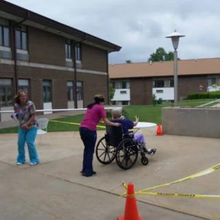 Two people assist a person in a wheelchair on an outdoor pathway near buildings.
