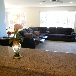 A cozy living room with a large sofa set, a vase of flowers on the counter, and sunlight streaming through the windows.