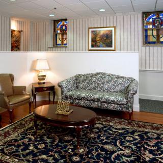 A cozy living room setup with a patterned sofa, armchair, and a wooden coffee table on an ornate rug. Stained glass windows and wall art enhance the decor.