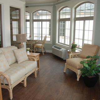 A cozy living room with ample natural light, featuring comfortable furniture, large windows, and indoor plants.