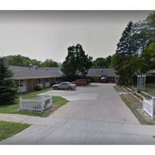A suburban residential area featuring single-story houses, a driveway, parked cars, and surrounding trees.