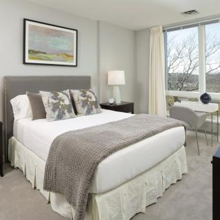 A modern bedroom featuring a neatly made bed with neutral-colored bedding and decorative pillows, complemented by a bedside lamp, window view, and a framed painting.
