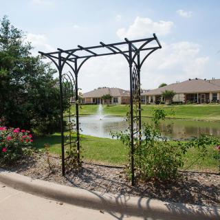 A decorative metal archway stands in a garden overlooking a pond with fountains and residential homes in the background.