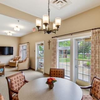 A cozy living room with a dining area, featuring a chandelier, comfortable chairs, and large windows overlooking a garden.