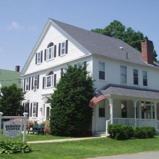 A traditional white house with a porch, surrounded by greenery and displaying flags.