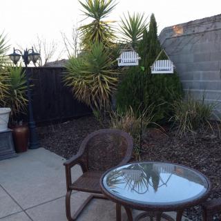 A cozy patio with a glass-top table and wicker chair, surrounded by lush greenery.