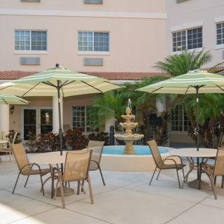 A patio area with tables and chairs shaded by umbrellas, set near a fountain and surrounded by palm trees.