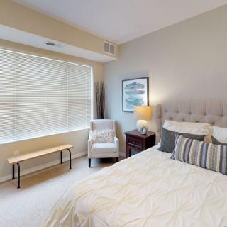 A cozy and well-lit bedroom with neutral colors, featuring a large window, a comfortable bed, an armchair, and tasteful decor.