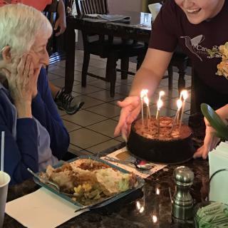 An elderly person smiling as a birthday cake with lit candles is presented in a restaurant setting.