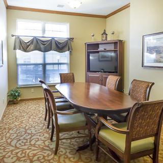 A small conference room with an oval wooden table surrounded by chairs, featuring a television, window with drapes, and patterned carpet.