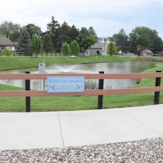 A serene view of a park featuring a pond surrounded by greenery and a wooden fence.