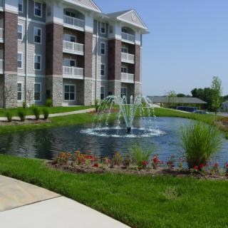 A modern apartment building with a landscaped pond and fountain in the foreground.
