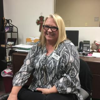 A woman in a patterned blouse smiles while seated in an office environment.