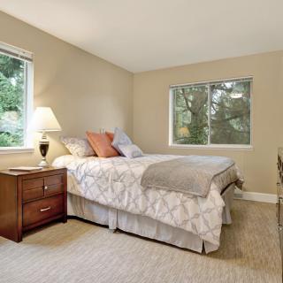 A cozy bedroom with a neatly made bed, two windows, and a nightstand with a lamp.