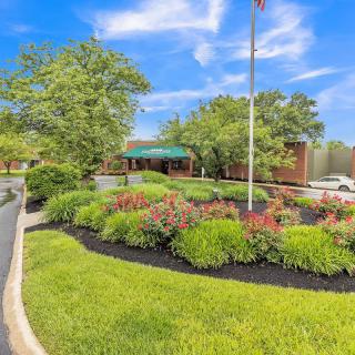 A beautifully landscaped area with a flagpole and a building in the background, surrounded by lush greenery and colorful flowers under a bright blue sky.