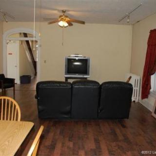 A cozy living room with a television, sofa, dining table, wooden flooring, and a ceiling fan.