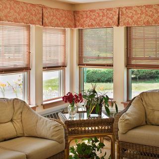 A cozy sunroom with comfortable armchairs and a table, surrounded by large windows with blinds filtering natural light.