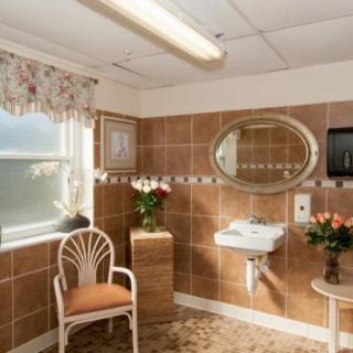 A cozy bathroom interior featuring brown tiles, a round mirror, a chair, and decorative flowers.